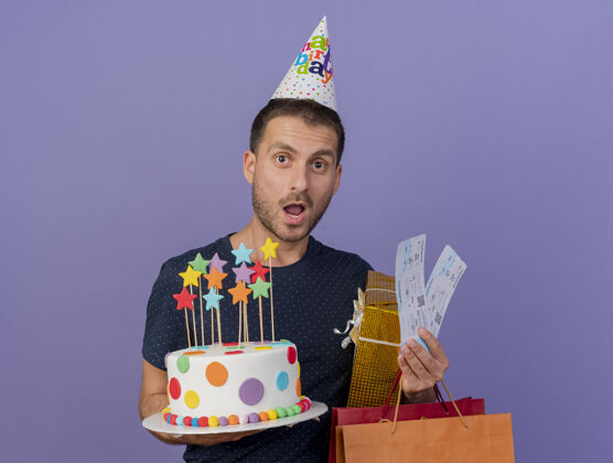盒子戴着生日帽的帅哥高加索人拿着生日蛋糕纸购物袋礼盒和机票 紫色背景上有复印空间包票男人
