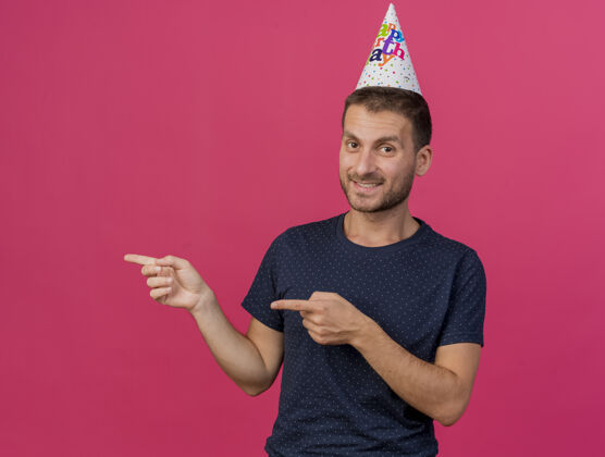 复制笑容可掬的白人帅哥戴着生日帽指着侧面 两手分开放在粉色背景上 留有复印空间两个指向男人