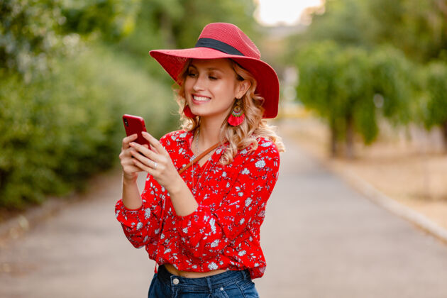 外表迷人的时尚金发微笑的女人在稻草红帽子和衬衫夏季时尚套装使用手机配饰魅力时尚