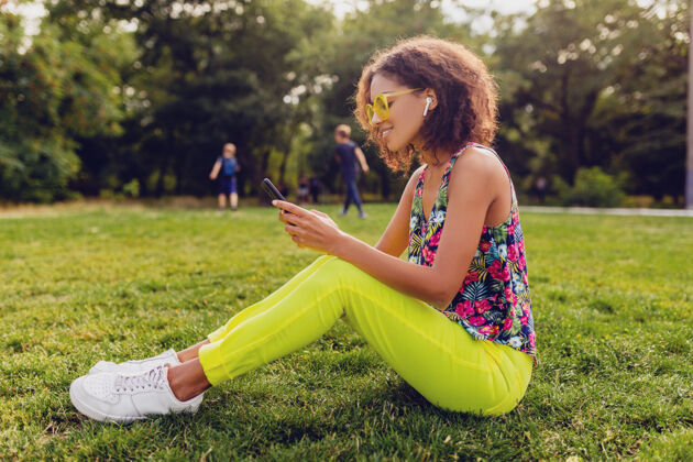 耳机年轻时尚微笑的黑人女子用智能手机听音乐无线耳机在公园里玩得开心 夏日时尚缤纷风格多彩女人人
