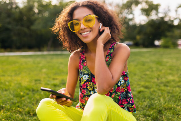 眼镜年轻时尚微笑的黑人女子使用智能手机 通过无线耳机听音乐 在公园里玩得很开心黄色音乐电话