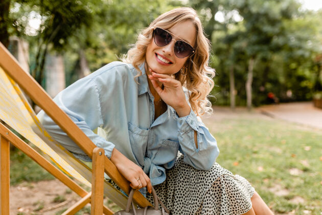 优雅迷人的金发微笑的女人坐在躺椅上穿着夏天的衣服时尚快乐手提包