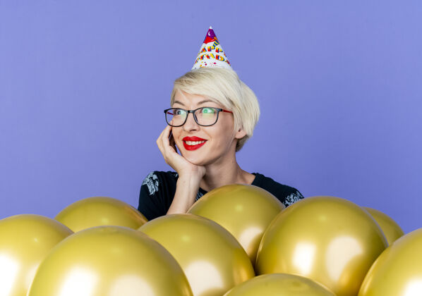 派对微笑的金发派对女郎戴着眼镜和生日帽站在气球后面把手放在下巴下面看着紫色背景上孤立的一面微笑女孩站着