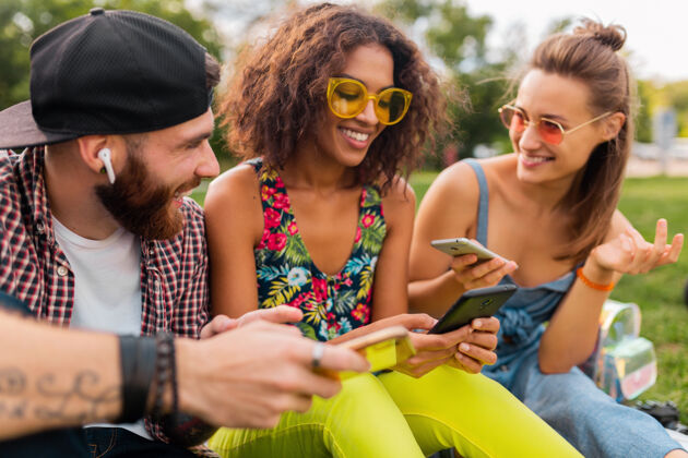 太阳镜快乐的年轻朋友们坐在公园里用智能手机微笑 男人和女人一起玩微笑团体音乐