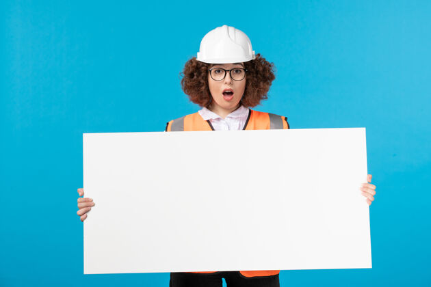 女性建设者穿着制服的女建筑工人的正面图 白色的普通办公桌 蓝色的墙壁板子女性手持