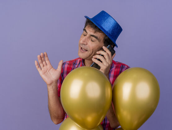 站着戴着派对帽的中年白种人站在气球后面讲电话 手举在空中 闭着眼睛孤立地站在紫色背景上眼睛气球中年