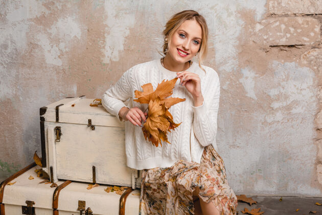 温暖迷人的微笑 穿着白色针织毛衣的时尚金发女人坐在手提箱上的肖像在街上的老式墙上季节美丽秋天