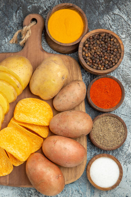 饮食木制砧板上的脆片和生土豆以及灰色桌子上不同香料的俯视图马铃薯开销可食用的水果