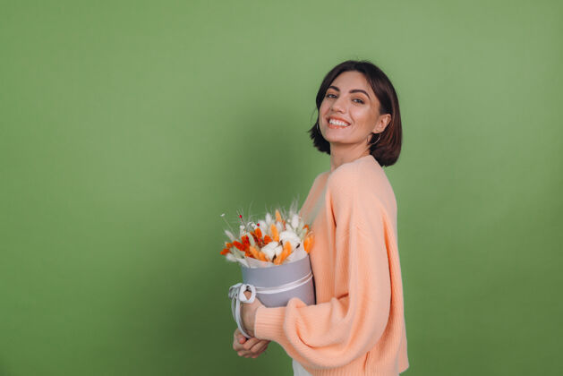 黑发年轻女子穿着休闲桃色毛衣隔离在绿橄榄墙上手持橙白色花盒组成的棉花 吉普赛拉小麦和拉古鲁斯作为礼物开心惊喜干花黑麦静物