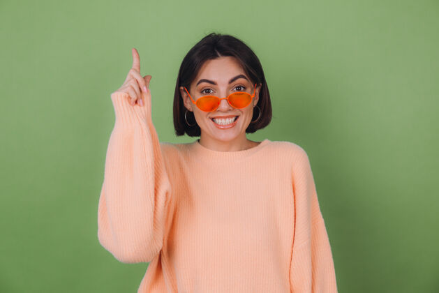 模特年轻时尚的女士穿着休闲的桃色毛衣 戴着橘黄色的眼镜 隔着绿橄榄色的墙壁兴奋地点着食指向上抄空间女性黑发点