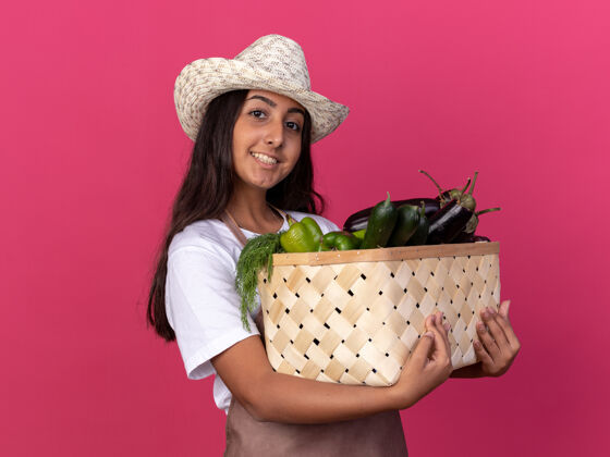 年轻快乐的年轻园丁女孩 围着围裙 戴着夏帽 手里拿着装满蔬菜的箱子 脸上带着微笑站在粉色的墙上微笑围裙立场