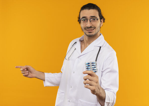 黄色高兴的年轻医生戴着医用眼镜 穿着医用长袍 手持听诊器和药片 指着旁边请男人年轻