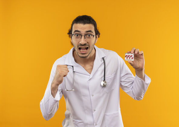 年轻人快乐的年轻医生戴着医用眼镜 穿着带听诊器的医用长袍 拿着药片 做着“是”的手势是的黄色药丸
