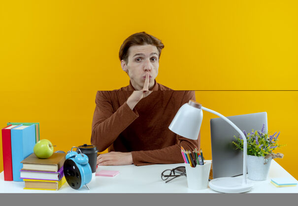 黄色一个年轻的学生男孩坐在书桌旁 手里拿着学习用具 做着沉默的手势工具男孩学生