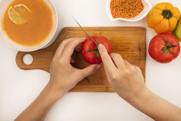 健康俯视图中的女性手切新鲜的西红柿在一个木制的菜板上用刀与扁豆汤在一个碗五颜六色的辣椒隔离在一个白色的表面五颜六色西红柿营养