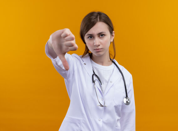 长袍年轻的女医生穿着医用长袍和听诊器 大拇指朝下放在隔离的橙色墙上 留有复印空间穿医学不愉快
