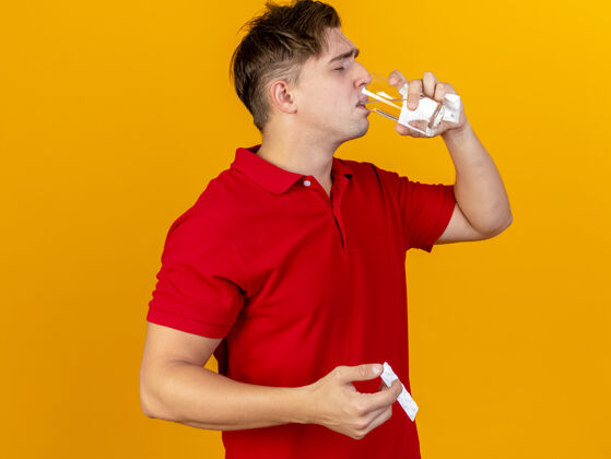 手势年轻英俊的金发病男子拿着餐巾纸吃药喝水从玻璃隔离在橙色的墙上服用玻璃快递