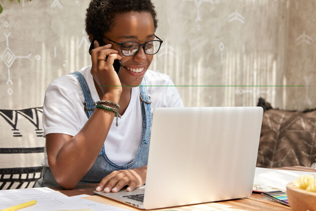专业时尚的非裔美国女人打电话智能手机看笔记本电脑 更新软件坐着自由职业者人类