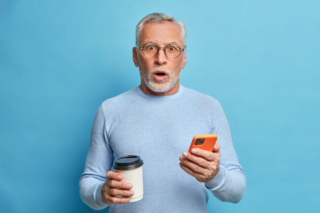 印象惊喜交集的成熟男人惊愕得喘不过气来拿着智能手机看新闻喝咖啡去接突发新闻穿休闲套头衫隔着蓝色的墙惊人老年人科技