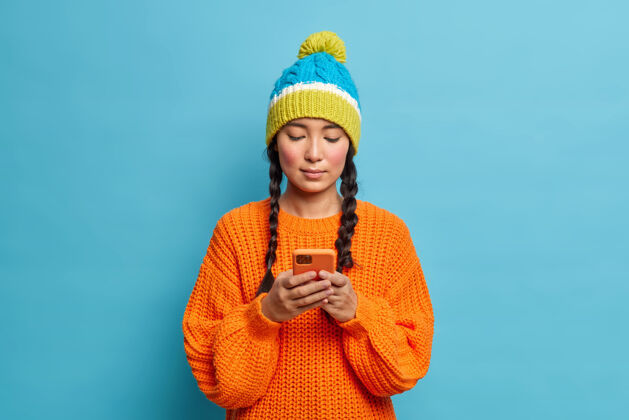 头发严肃的千禧一代女孩 辫子集中在智能手机显示屏上 使用无线网络 穿着时尚的冬装 安排与隔离在蓝色墙上的朋友在线聊天帽子黑发可爱