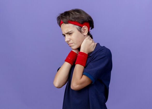 年轻疼痛的年轻英俊的运动男孩戴着头带和戴着牙套的腕带站在侧视图中俯视 手放在脖子后面 隔离在紫色的墙上 留有复制空间保持颈部头带