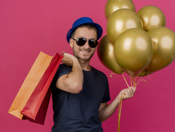 购物面带微笑的白种帅哥 戴着太阳眼镜 头戴蓝色派对帽 手里拿着氦气球和纸购物袋 粉色背景上有复印空间氦眼镜包