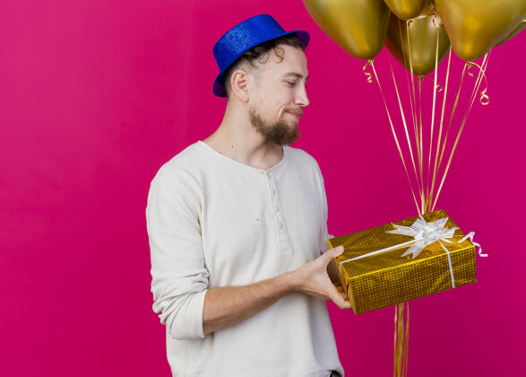 盒子高兴的年轻英俊的斯拉夫党家伙戴着党的帽子拿着气球和礼品盒看着礼品盒上的深红色背景与复制空间隔离年轻人拜托礼物