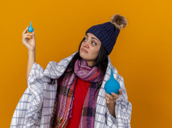 小体贴的年轻白种人生病的女孩戴着冬天的帽子和围巾包裹在格子布里拿着灌肠剂看着一个孤立的小家伙在橙色的背景下复制空间女孩帽子疾病