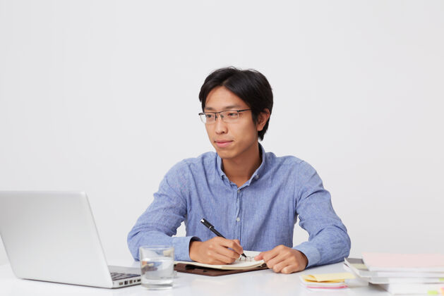 认真一位戴眼镜的亚洲年轻富商 面带沉思 在笔记本上写字 在桌旁工作 笔记本电脑盖在白墙上计划桌子亚洲人