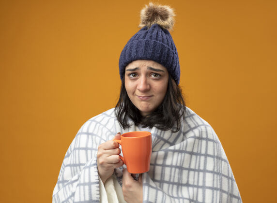 格子虚弱的年轻白种人生病的女孩穿着长袍 戴着格子呢的冬帽 手里拿着一杯茶 看着孤立在橙色背景上的相机相机女孩长袍
