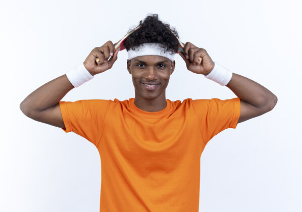 运动微笑着的年轻的非裔美国人 戴着头带和腕带 头上戴着乒乓球拍 隔离在白墙上盖年轻男人