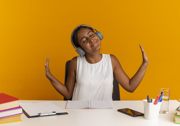 手年轻的女学生高兴地闭上眼睛 拿着学习用具坐在书桌旁 戴着耳机听音乐 双手分开放在橙色的墙上耳机桌子坐着