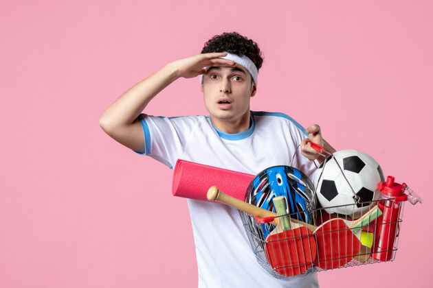 前面正面图穿着运动服的年轻男子 带着装满运动用品的篮子 粉色的墙壁健身房体育事物