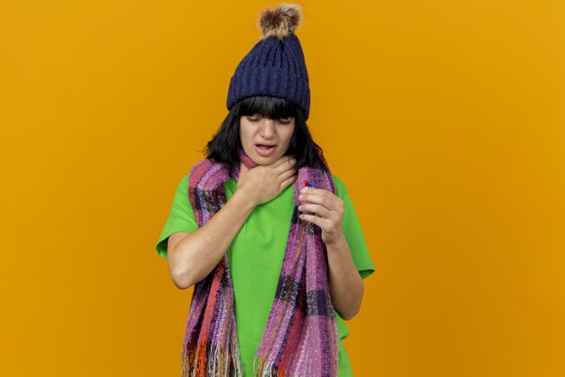 持有疼痛的年轻白种人女孩戴着冬天的帽子和围巾拿着胶囊手放在喉咙上闭着眼睛隔离在橙色的背景和复制空间围巾胶囊疾病