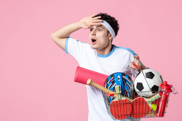 粉红色正面图穿着运动服的年轻男子 带着装满运动用品的篮子 粉色的墙壁充分体育东西