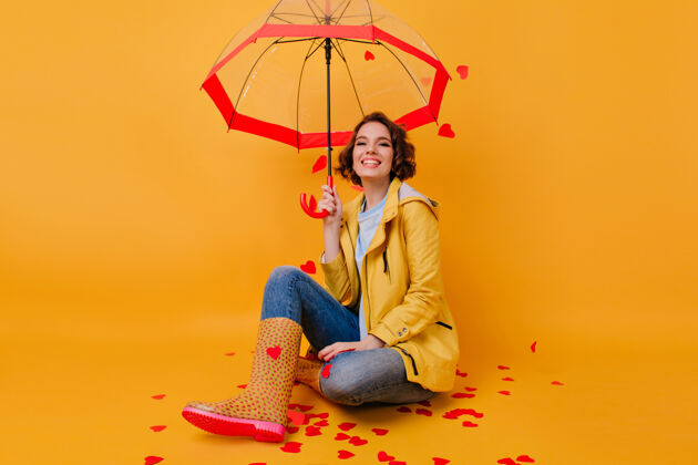伞穿着滑稽胶鞋的梦幻女孩双腿交叉坐在黄色的墙上穿着鲜艳夹克的浪漫女人与阳伞合影的室内肖像魅力季节年轻