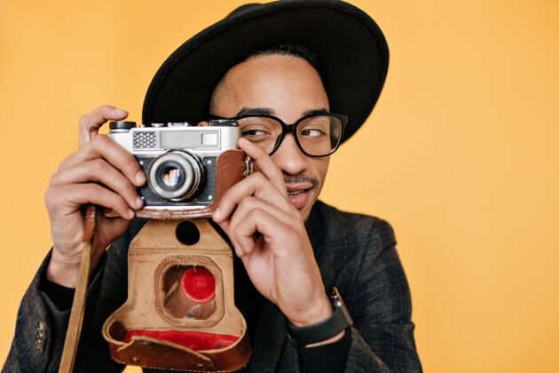 积极在黄色的墙上 一个非洲年轻人开玩笑地和相机合影一个戴着优雅帽子的天才摄影师的室内照片年轻人室内男人