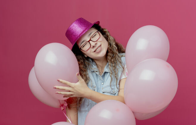 封闭闭上眼睛微笑着戴着眼镜和粉红色帽子的年轻女孩站在孤立的粉红色气球中间站着女孩微笑