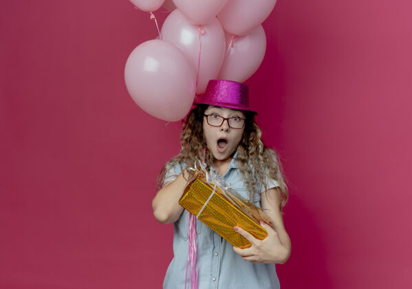 女孩惊讶的年轻女孩戴着眼镜和粉红色的帽子拿着礼盒和气球孤立在粉红色持有盒子帽子