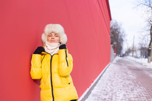 休闲在一个冬天阳光明媚的日子里 穿着暖和衣服的红墙背景上的快乐年轻女子夹克穿着墙