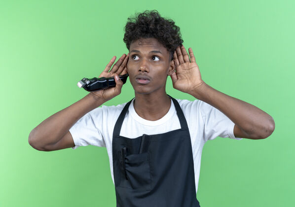 思考年轻的美国黑人男理发师穿着制服 拿着理发器 在绿色的墙上展示着孤立的倾听姿态手势显示壁板