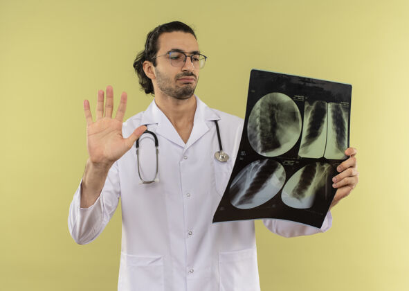 眼镜年轻的男医生戴着眼镜 穿着白色长袍 手持听诊器 看着x光片显示绿色的停止手势思考白色X光