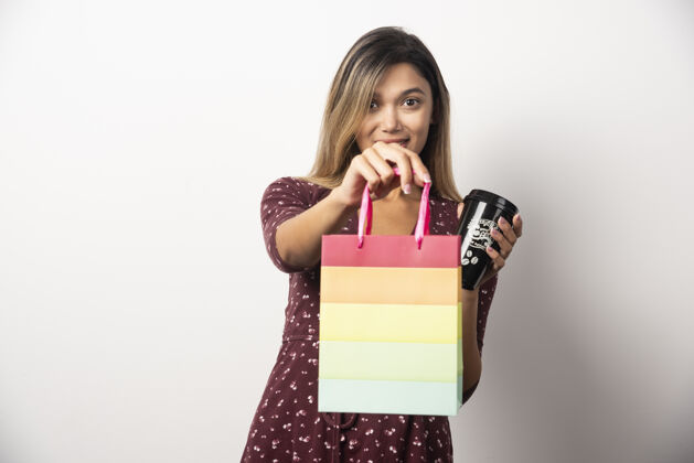 包年轻女子拿着一个小购物袋和一杯饮料在白墙上购物女性年轻
