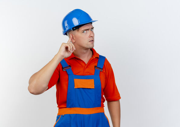 年轻严格的年轻男性建筑工人穿着制服和安全帽把手指放在耳朵上的白色头盔严格耳朵