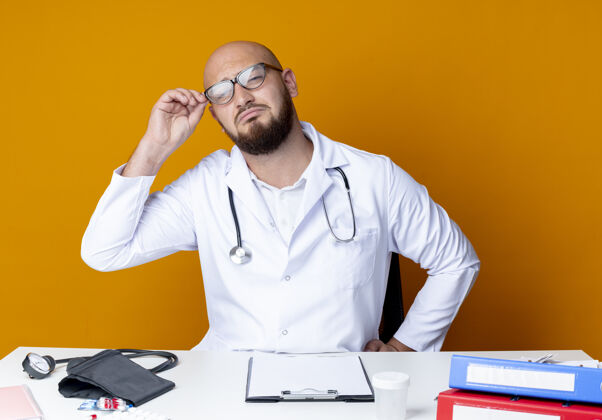 工作悲伤的年轻秃头男医生穿着医用长袍和听诊器坐在工作台上拿着医疗工具戴上橙色的眼镜秃头长袍年轻