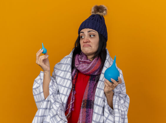 人困惑的年轻生病的女人戴着冬天的帽子 围巾裹着格子布 拿着灌肠剂 看着隔离在橙色墙上的小家伙疾病冬天帽子