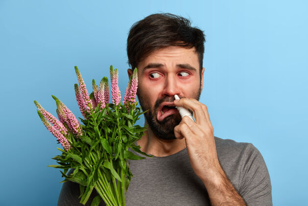 治疗不快乐的男人感觉不舒服 对花粉过敏 对植物过敏 用鼻喷雾剂喷鼻 需要治疗 在蓝色墙壁上摆姿势 治疗鼻炎医学概念草药室内年轻