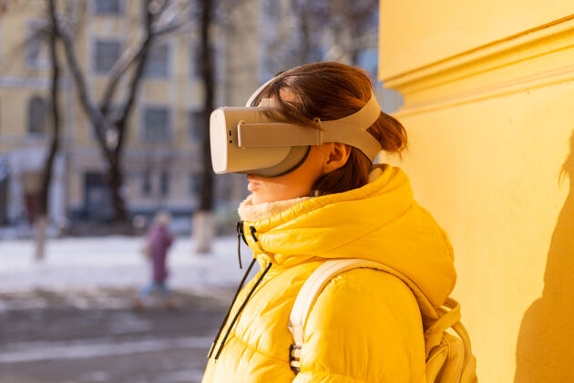 可穿戴一个戴着虚拟现实眼镜的女人的肖像 在冬天的阳光下 穿着暖和的衣服 靠在黄色的墙上耳机现实创新