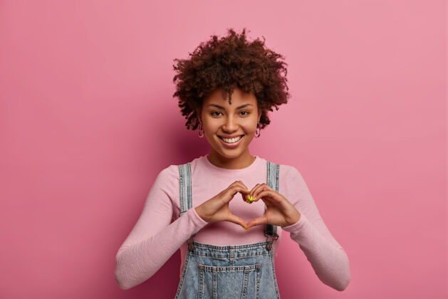 罗西开朗的非洲裔美国女人用手做心形 承认爱情 积极微笑 穿着休闲装 对着玫瑰色的粉彩墙摆姿势浪漫的感觉 肢体语言的概念室内形状模特