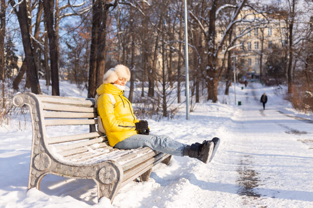 外面冬天的快乐年轻女子 穿着暖和的衣服 在阳光明媚的日子里 坐在长椅上 独自享受着新鲜的空气和咖啡微笑酷长凳